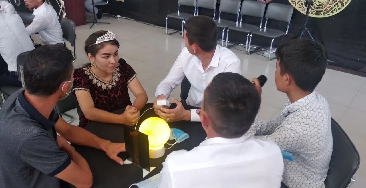 Stol atrofida o'tirgan jamoa a'zolari, o'rtada sariq chiroq yonib turibdi